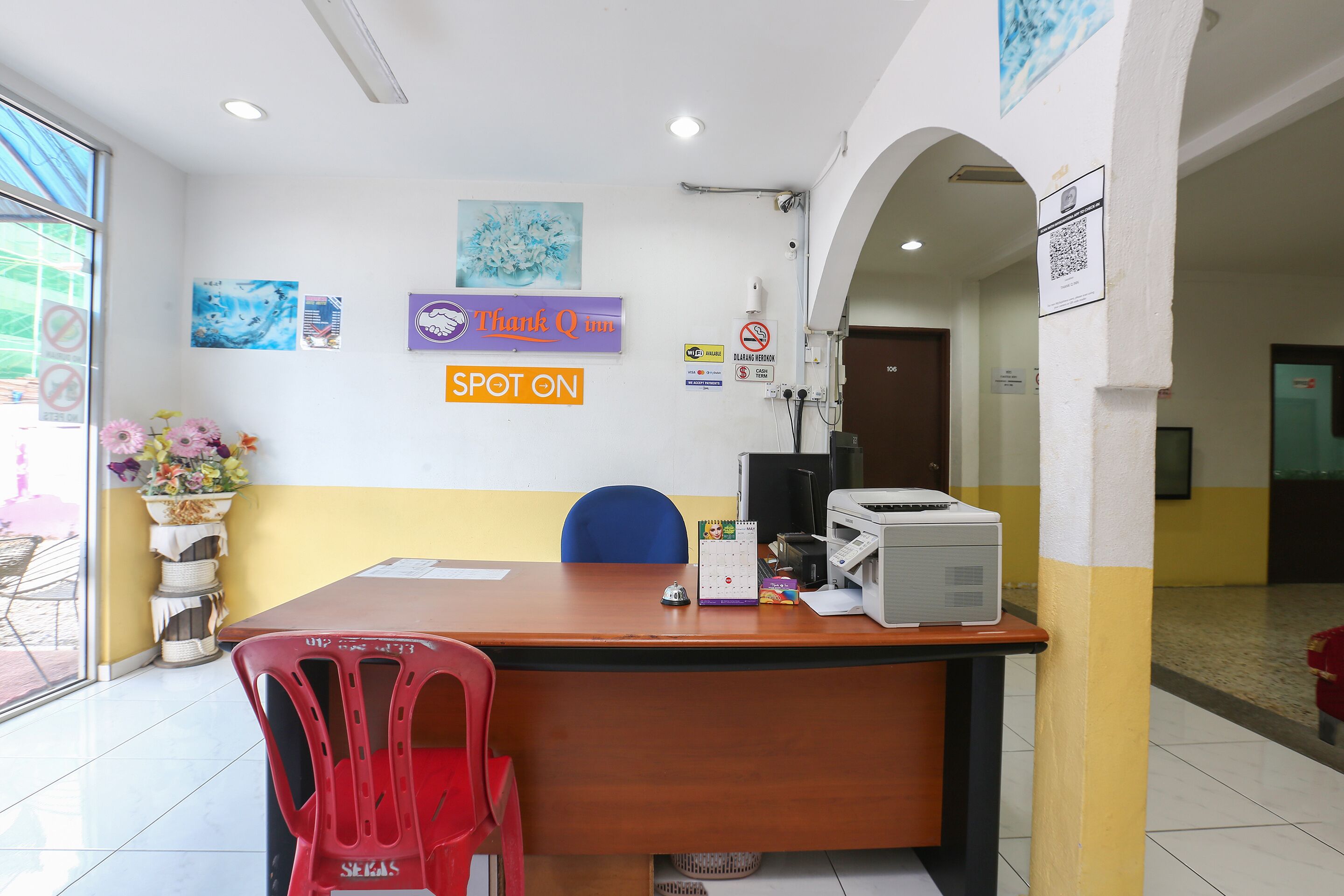 A reception area with a wooden desk, a red plastic chair, and a vase of flowers. There are wall decorations and a calendar on the wall.