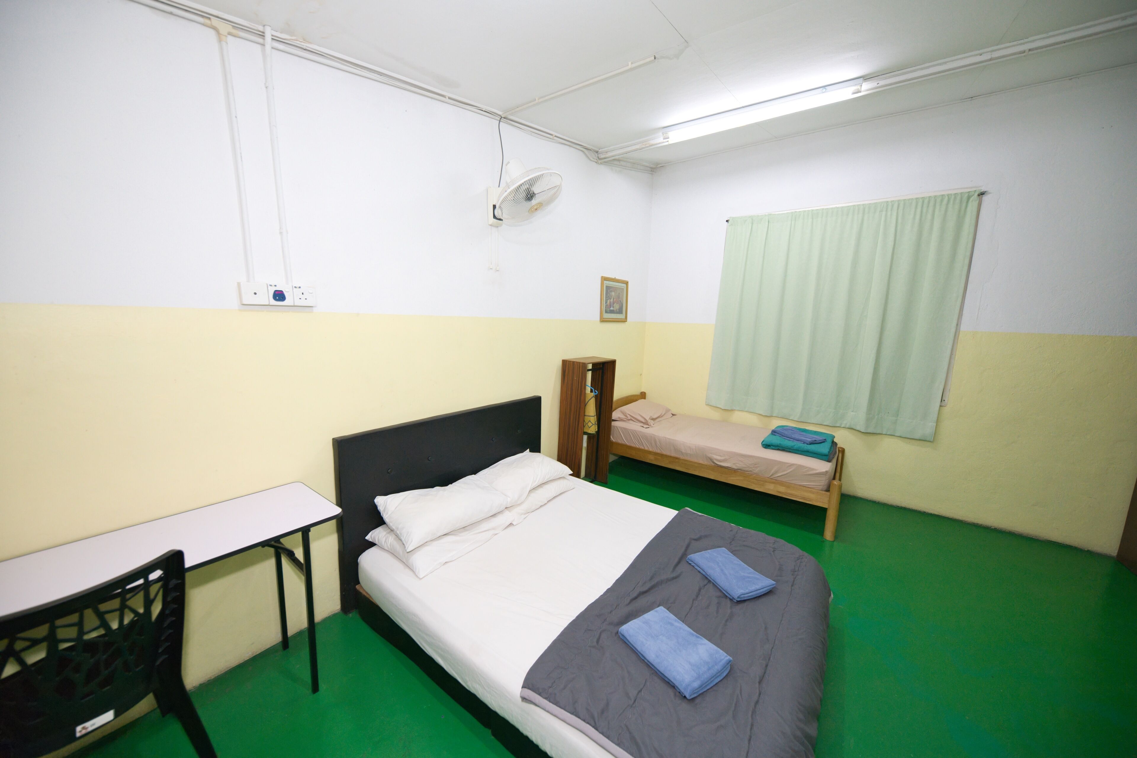 A small, single-room accommodation with a bed, a desk, a chair, a bedside table, a fan, and a window with curtains.