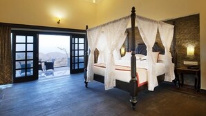 A four-poster bed with a canopy, a stone wall, and a view of mountains through a glass door. - The Wild Retreat (Kumbhalgarh)