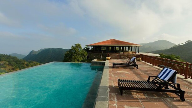 A pool area with a clear view of the mountains, a covered seating area, and a lounge chair. - The Wild Retreat (Kumbhalgarh)