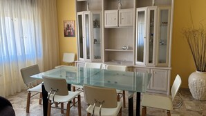 Un comedor con una mesa de vidrio y sillas blancas, un armario blanco con puertas de cristal, y un jarrón con plantas secas.