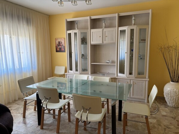 Un comedor con una mesa de vidrio y sillas blancas, un armario blanco con puertas de cristal, y un jarrón con plantas secas.
