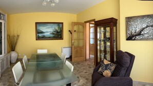 Un comedor con una mesa de vidrio, sillas blancas, un mueble con puertas de cristal, un cuadro y una puerta que da a otra habitación.