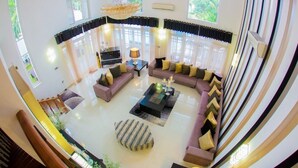 Ein großzügiger Wohnbereich mit einer großen Sofagarnitur, einem Couchtisch und einem gemusterten Hocker. Eine Chandelier hängt von der Decke und ein großes Fenster mit durchsichtigen Vorhängen.