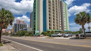 Ein hohes Hotel mit einer grün-beigen Fassade, mehreren Balkonen und einem Parkplatz mit Autos.