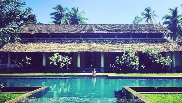 Piscina com uma pessoa na água, rodeada por um edifício com telhado em azulejos e palmeiras ao fundo.
