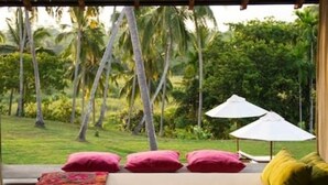 Quarto com cama, almofadas e vista para um jardim exuberante com palmeiras.