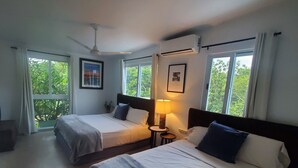 A hotel room with two beds, a ceiling fan, an air conditioning unit, a small table with a lamp, and large windows with curtains.