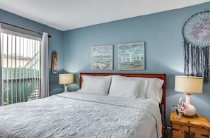 A bedroom with a large bed, two wall art pieces depicting boats, a dreamcatcher, and a nightstand with a lamp and flowers.
