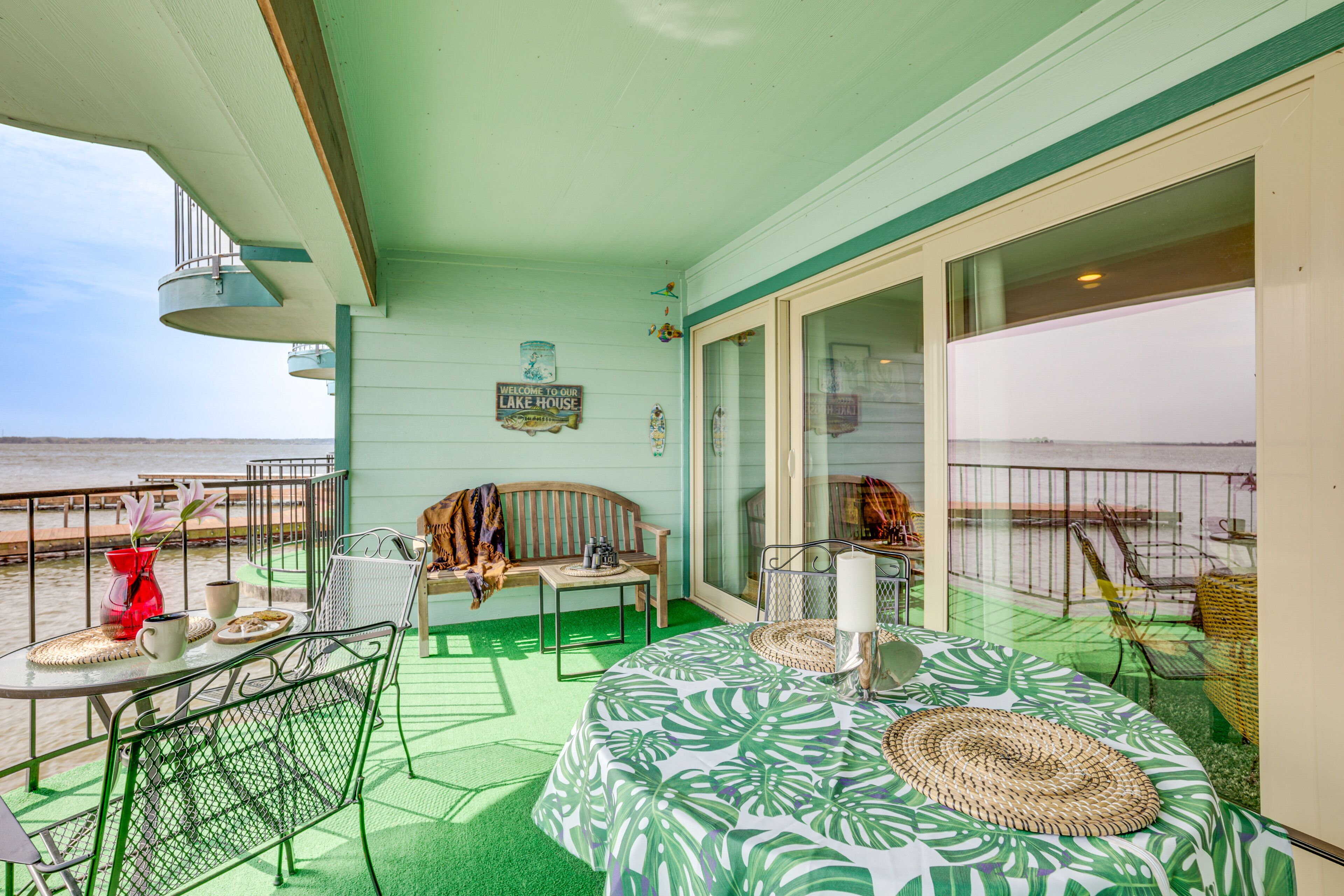 A balcony with a view of the beach, a table with a woven placemat, a bench, and a sign reading "Welcome to our Lake House".
