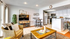 Ein modernes Wohnzimmer mit Sofa, Couchtisch und Fernseher. Es beinhaltet eine Küche mit weißen Schränken, einem Kühlschrank und einer Mikrowelle.