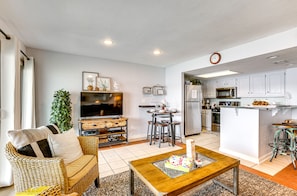 A modern living room with a sofa, coffee table, and a TV. It includes a kitchen with white cabinets, a refrigerator, and a microwave.