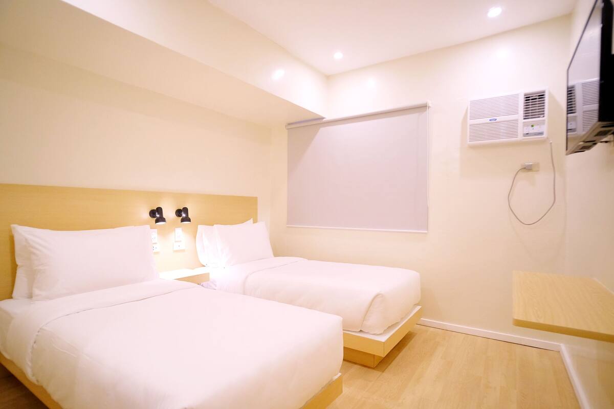 A hotel room with two beds, a bedside table, a wall-mounted air conditioner, and a window with a blind.