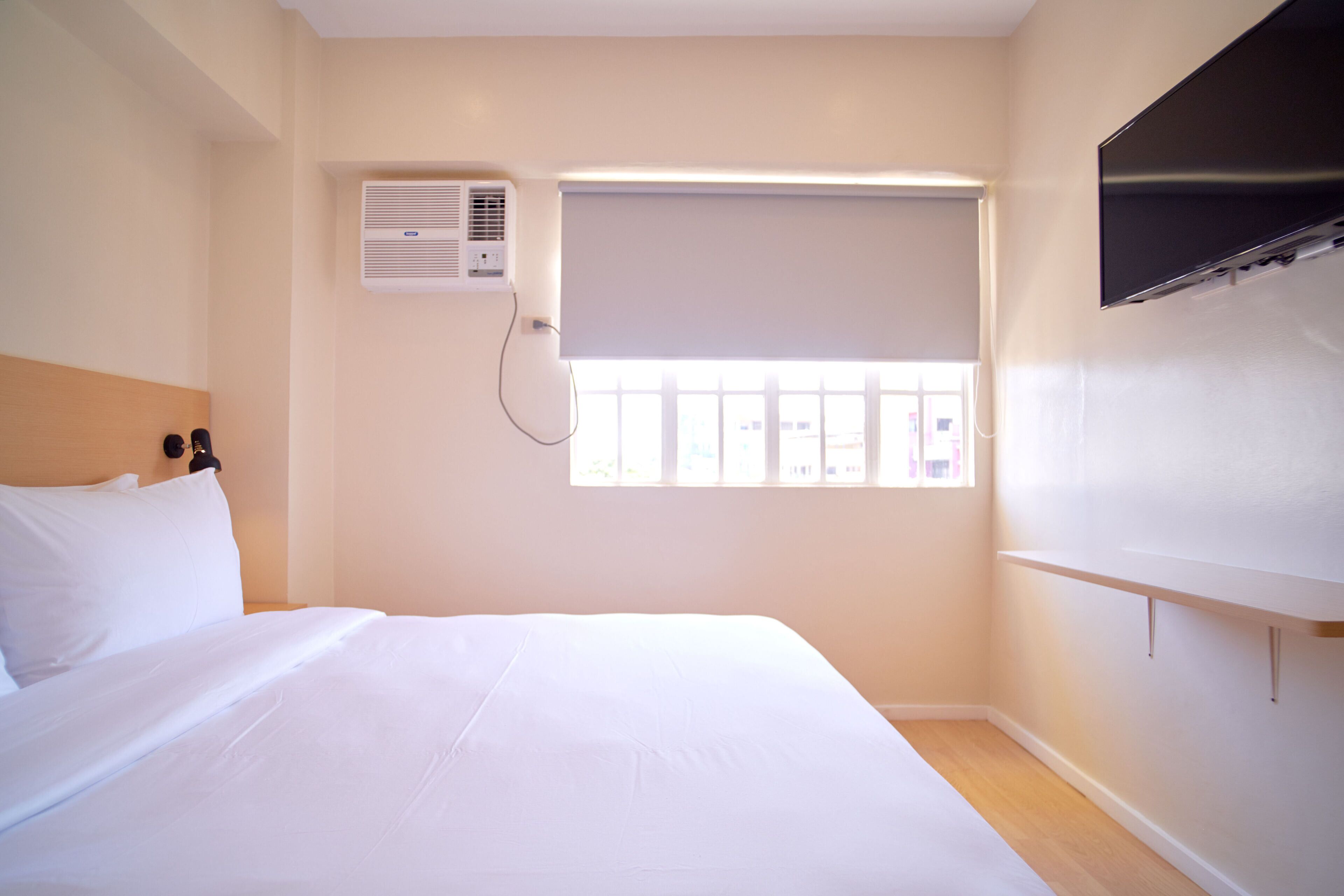 A hotel room with a bed, a window with blinds, an air conditioning unit, and a mounted TV.