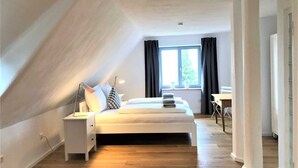 Un dormitorio con techo inclinado, una cama con almohadas, una mesita de noche, un escritorio y una ventana con cortinas.