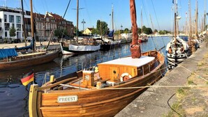 Un barco de madera amarrado en un puerto deportivo, con cielo despejado y edificios al fondo.