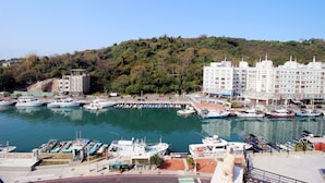 A marina with boats docked, a building with multiple floors, and a hillside with trees. - Hama Star (Kaohsiung)