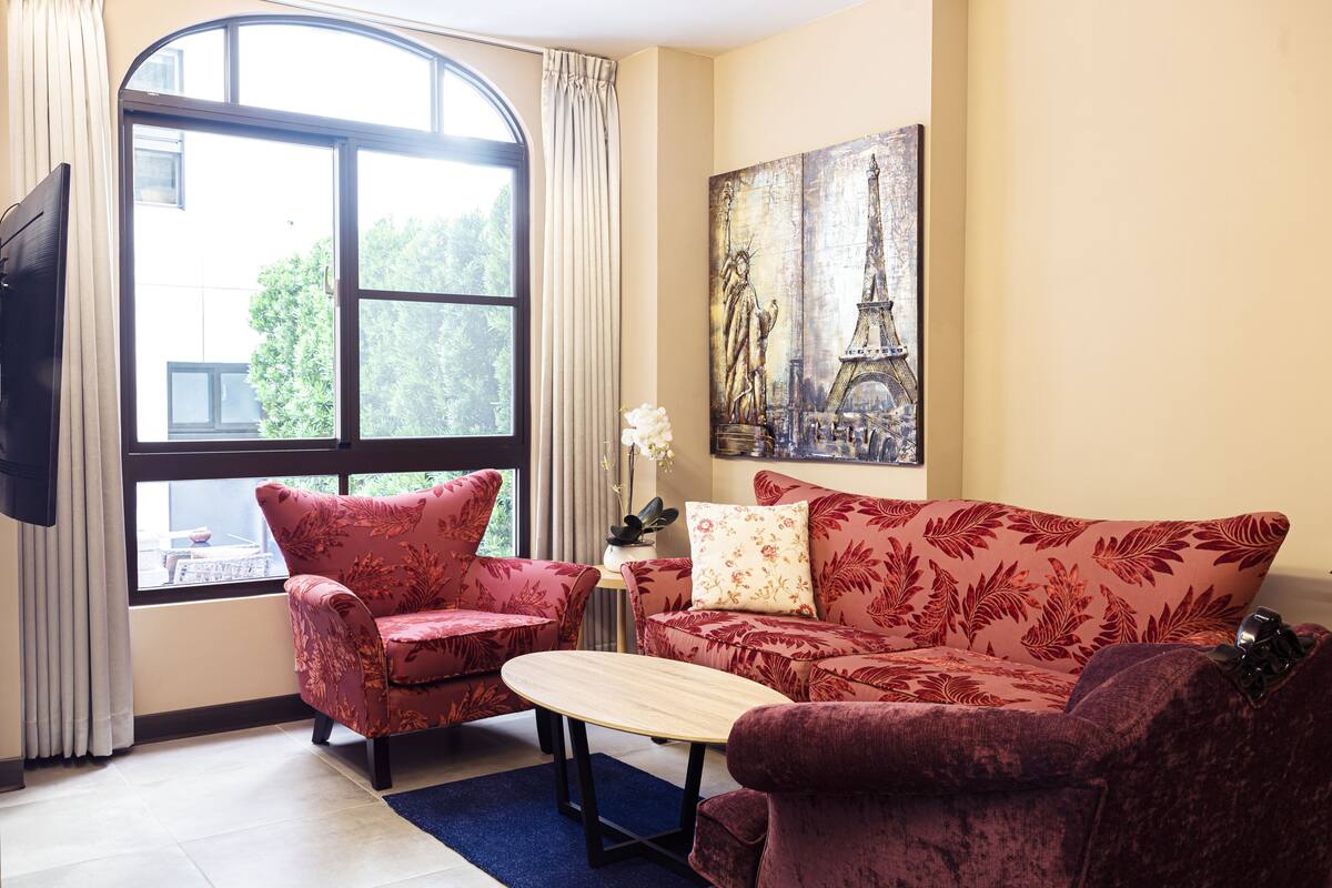 Uma sala de estar com um sofá, duas poltronas, uma mesa de centro, um quadro da Torre Eiffel e uma janela grande com cortinas.