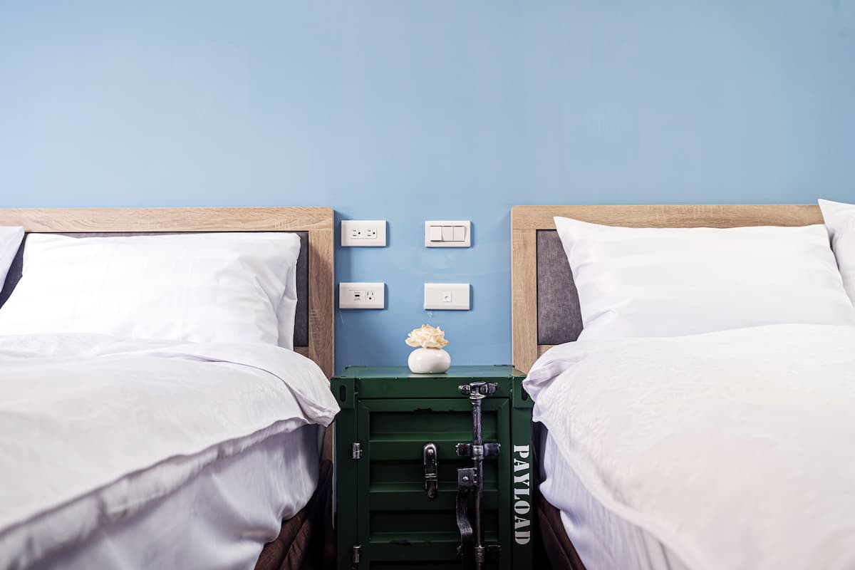 Quarto duplo com duas camas, um móvel verde para guardar bagagem, um vaso com flores e uma parede azul.
