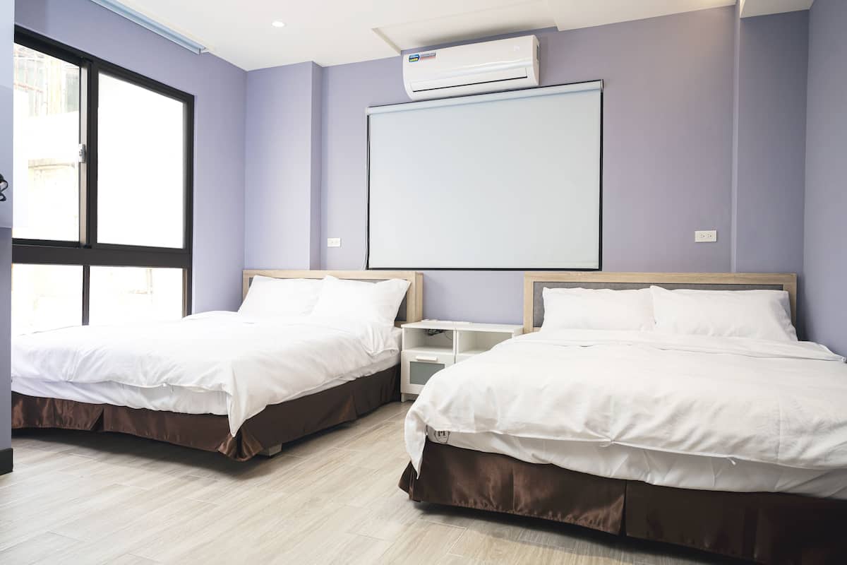 Quarto de hotel com duas camas, uma janela grande, ar condicionado e um quadro branco na parede.