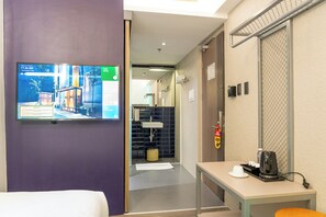 A hotel room with a television displaying room information, a bed, a table with a coffee maker, and a bathroom visible through an open door.