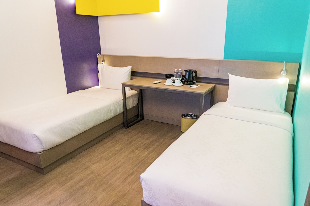 Ein Hotelzimmer mit zwei Einzelbetten, einem kleinen Tisch mit Wasserkocher und einem Mülleimer.