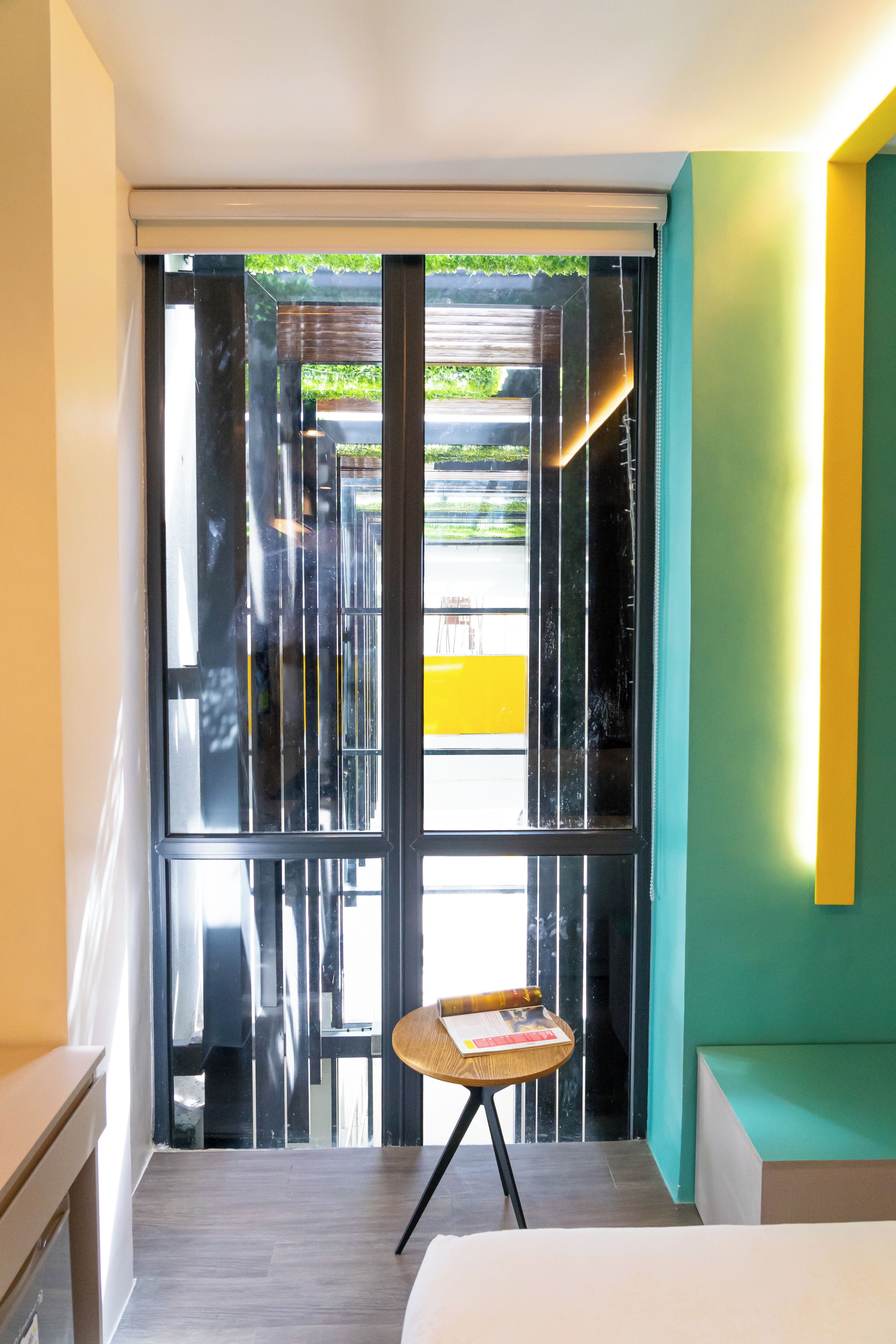 A hotel room with a bed, a small table with a book, and a view through a glass door.