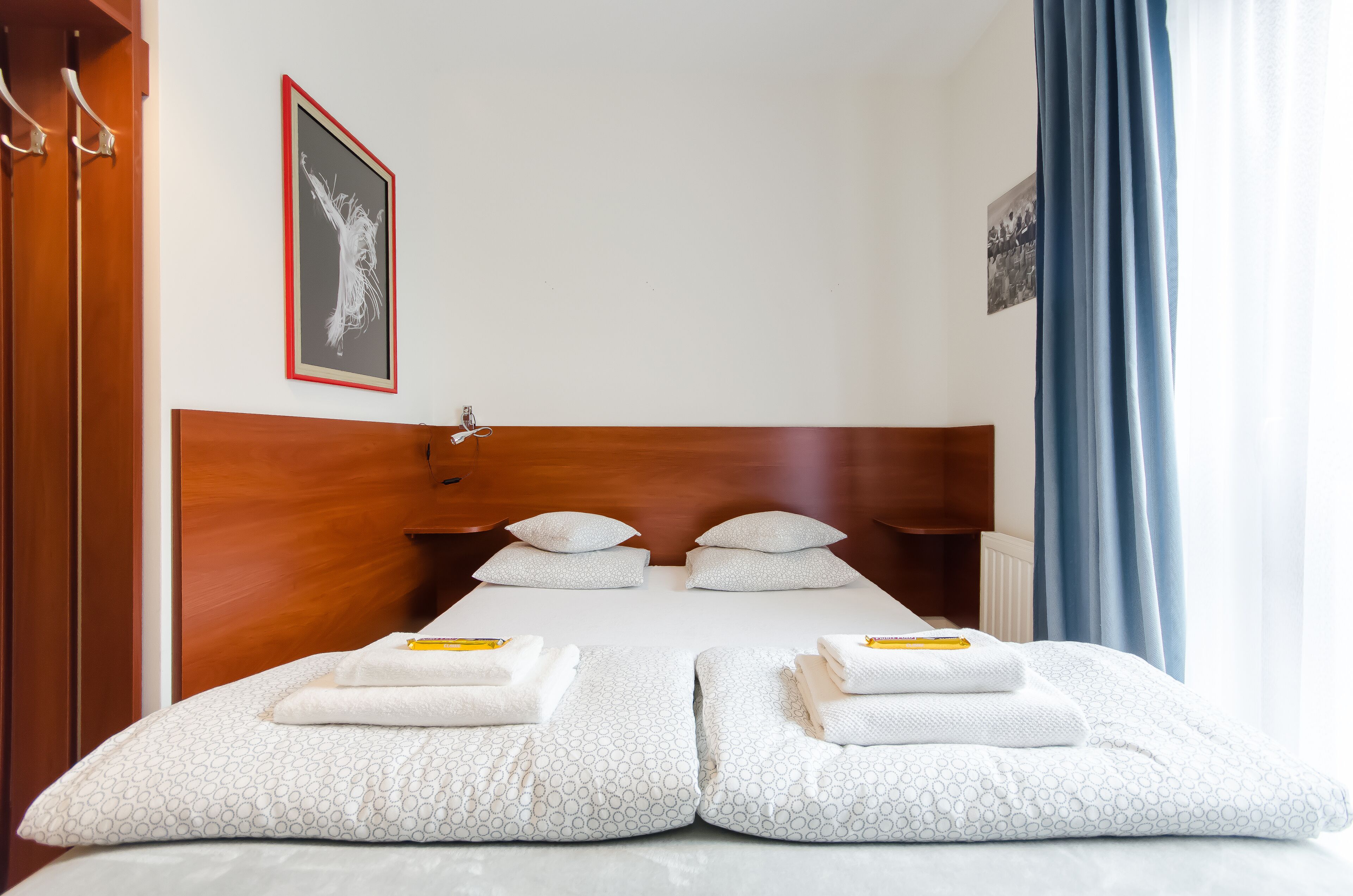 Ein Hotelzimmer mit einem hölzernen Kopfteil, zwei Betten mit weißer Bettwäsche und Handtüchern, einem gerahmten Bild an der Wand und einem Fenster mit blauen Vorhängen.