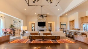 A spacious living area with a fireplace, a dining table with wicker chairs, and a kitchen in the background.