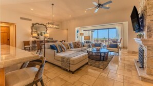 A spacious living room with a large sofa, a coffee table, and a dining area with a table and chairs. There is a fireplace and a large window offering a view of the outdoors.