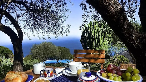 An outdoor dining setup with a table set for breakfast, including croissants, bowls of jam, and a fruit bowl, overlooking a scenic view of the sea and trees. - Baglio La Luna (San Vito Lo Capo)