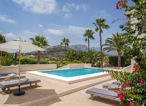 Un espace piscine agrémenté de chaises longues et de parasols, entouré de palmiers et dominé par un paysage montagneux.