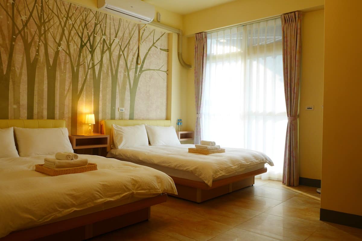 A hotel room with two beds, a large window with curtains, a wall with a tree mural, and a bedside table with a lamp.