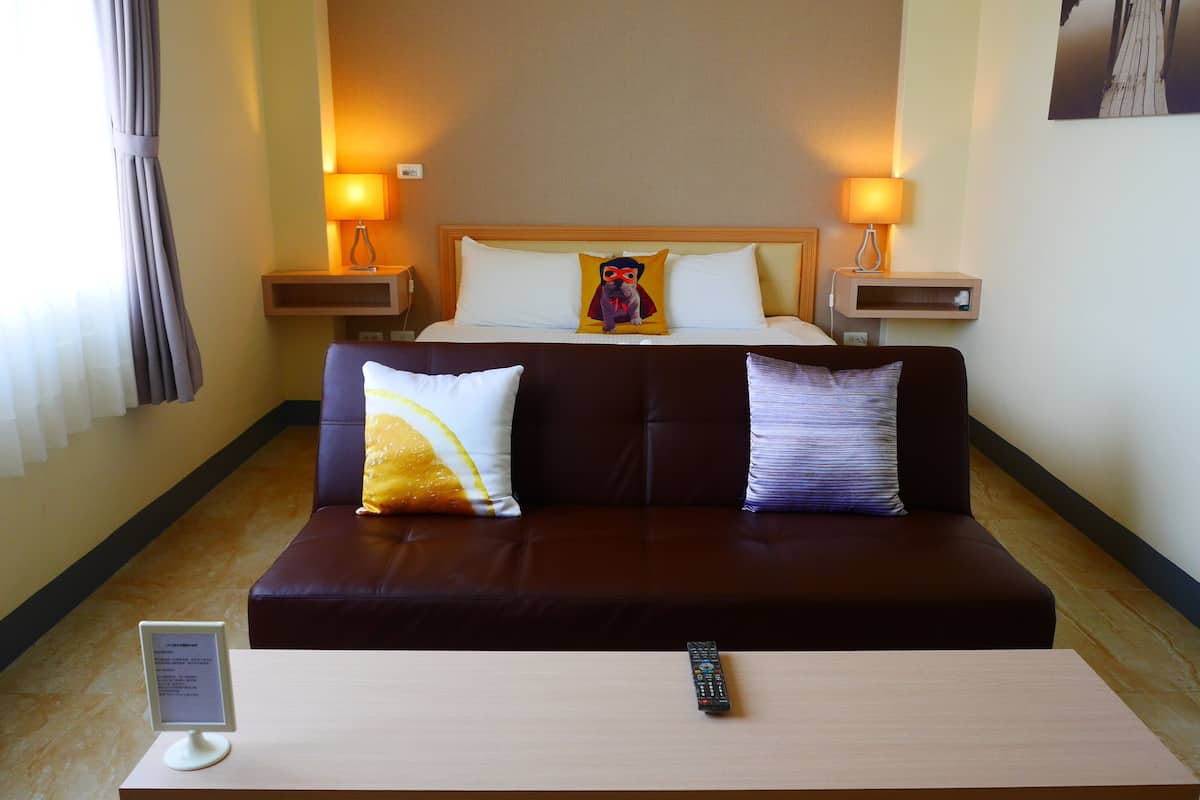 A hotel room with a bed, two nightstands, a sofa, a table, and a remote control.