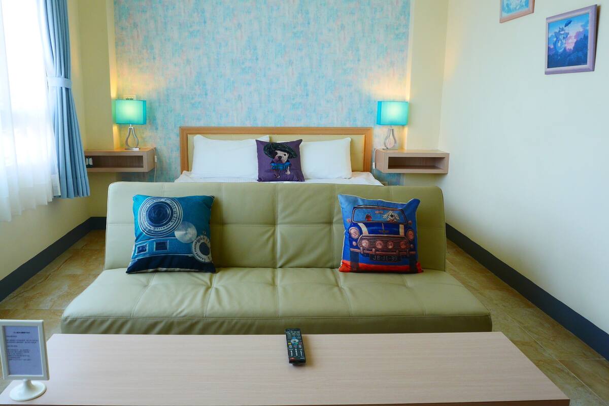 A hotel room with a green sofa, two pillows, a remote control on a table, and two bedside lamps.