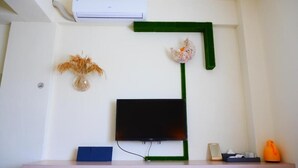 A modern room with a mounted flat-screen TV, a green pipe, a decorative plant, and a small table with a blue box and a white object. - Finest Hour Guesthouse (Chishang)