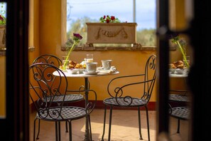 Un patio con sillas y mesas de metal, una pequeña mesa con pasteles y una taza, y vista a un edificio con una escultura.