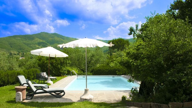 Una piscina con lettini e ombrelloni, circondata da vegetazione rigogliosa e colline.