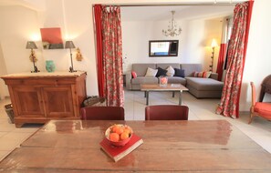 A dining area with a wooden table, chairs, and a bowl of fruit. There is a sofa and a television in the background. - L'anse Lympia (Nice)