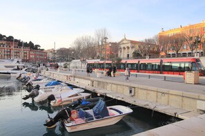 A marina with boats docked, a red tram, and buildings in the background. - L'anse Lympia (Nice)
