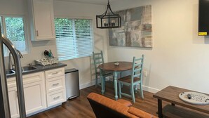 Cocina compacta con armarios blancos, un lavamanos de acero inoxidable y una zona de comedor con una mesa de madera y sillas color turquesa.