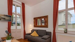 Ein Zimmer mit grauem Sofa, einem Holztisch, einer Topfpflanze, einem Fernseher und einem Fenster mit roten Vorhängen.