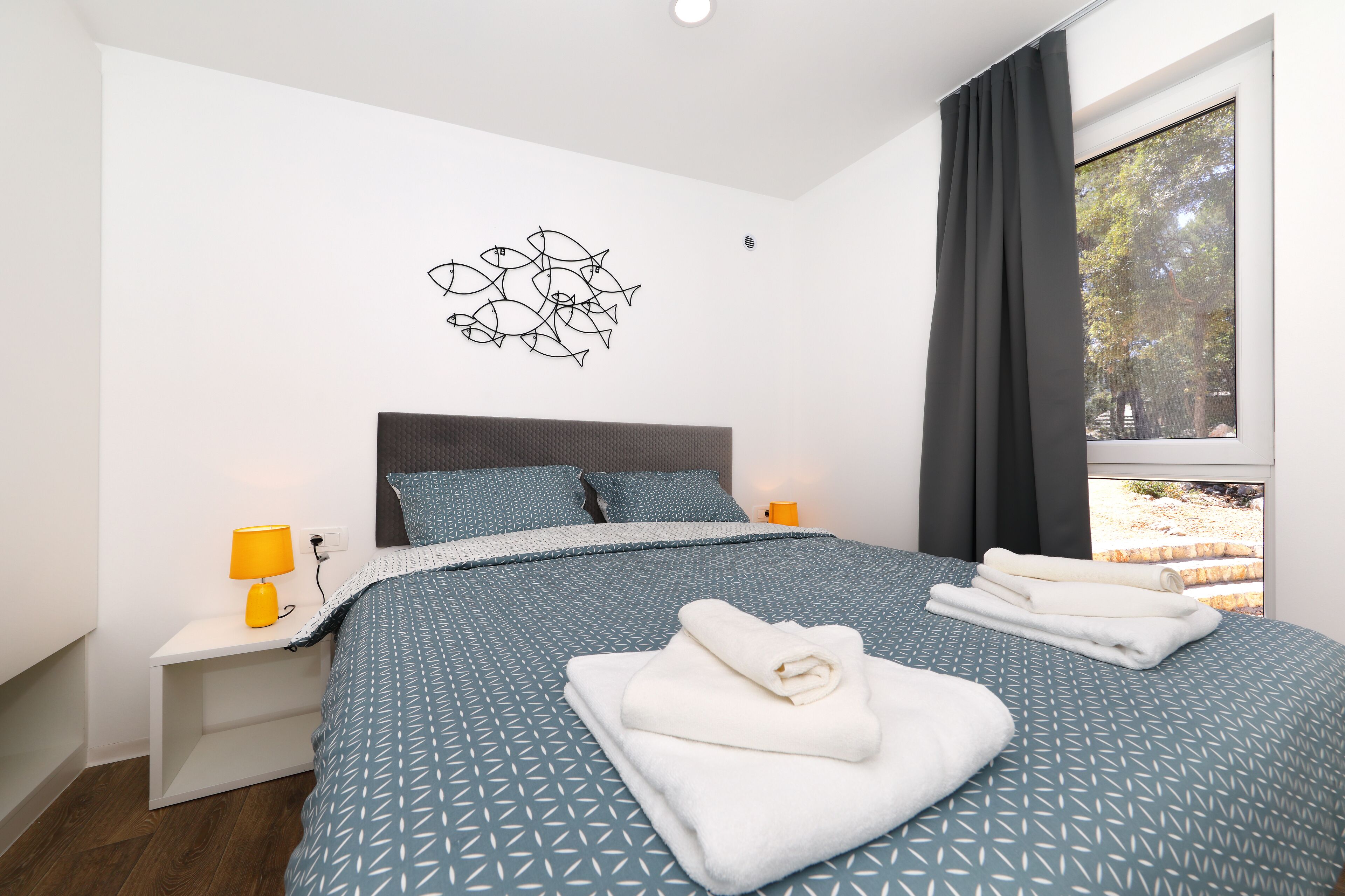 Quarto moderno com edredom estampado em padrão geométrico, cabeceira, duas mesas de cabeceira com abajures e vista para árvores pela janela.