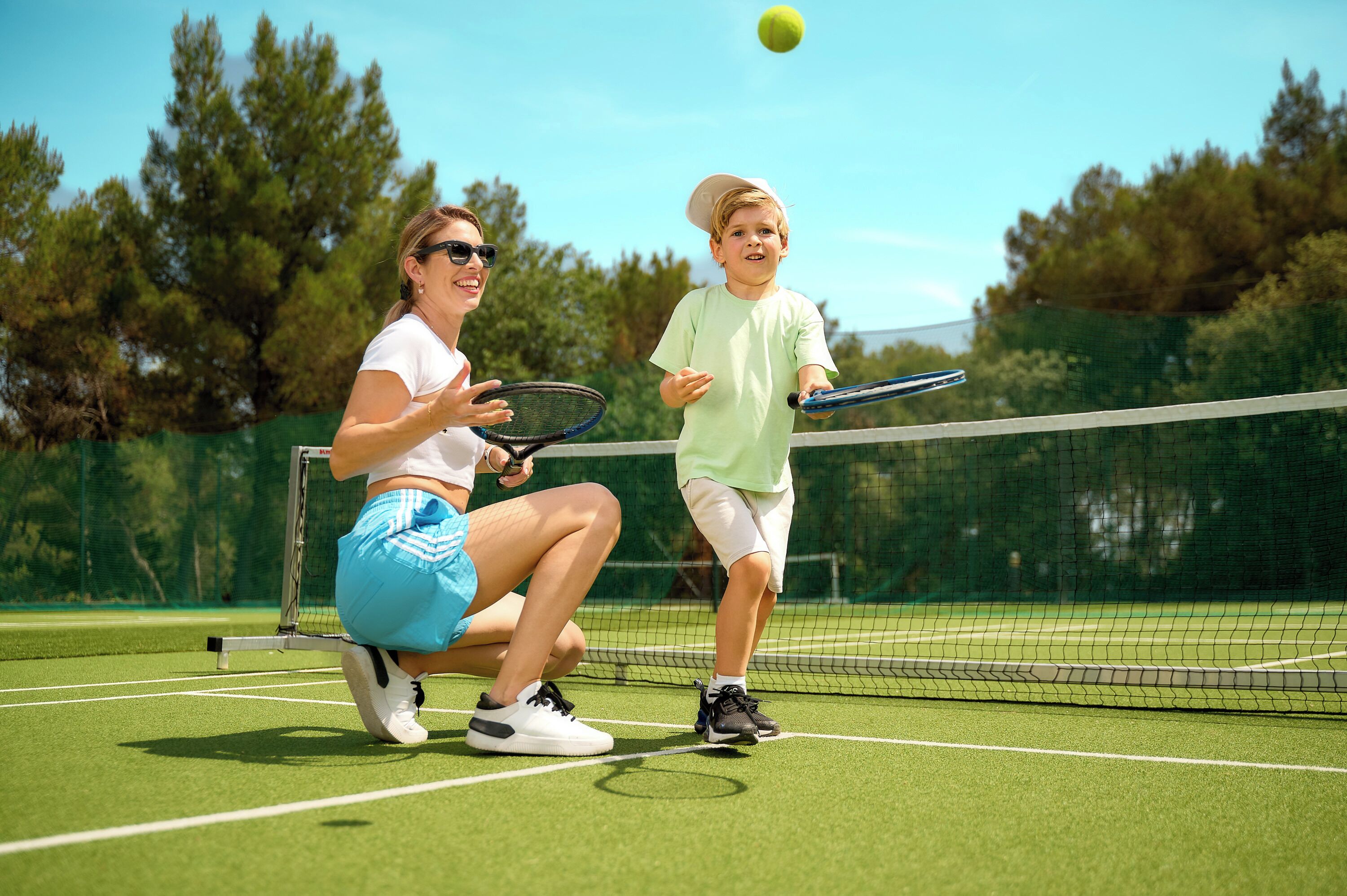 Un court de tennis avec un filet, une balle de tennis en l’air, et deux personnes, l’une assise et l’autre debout, tenant des raquettes de tennis.