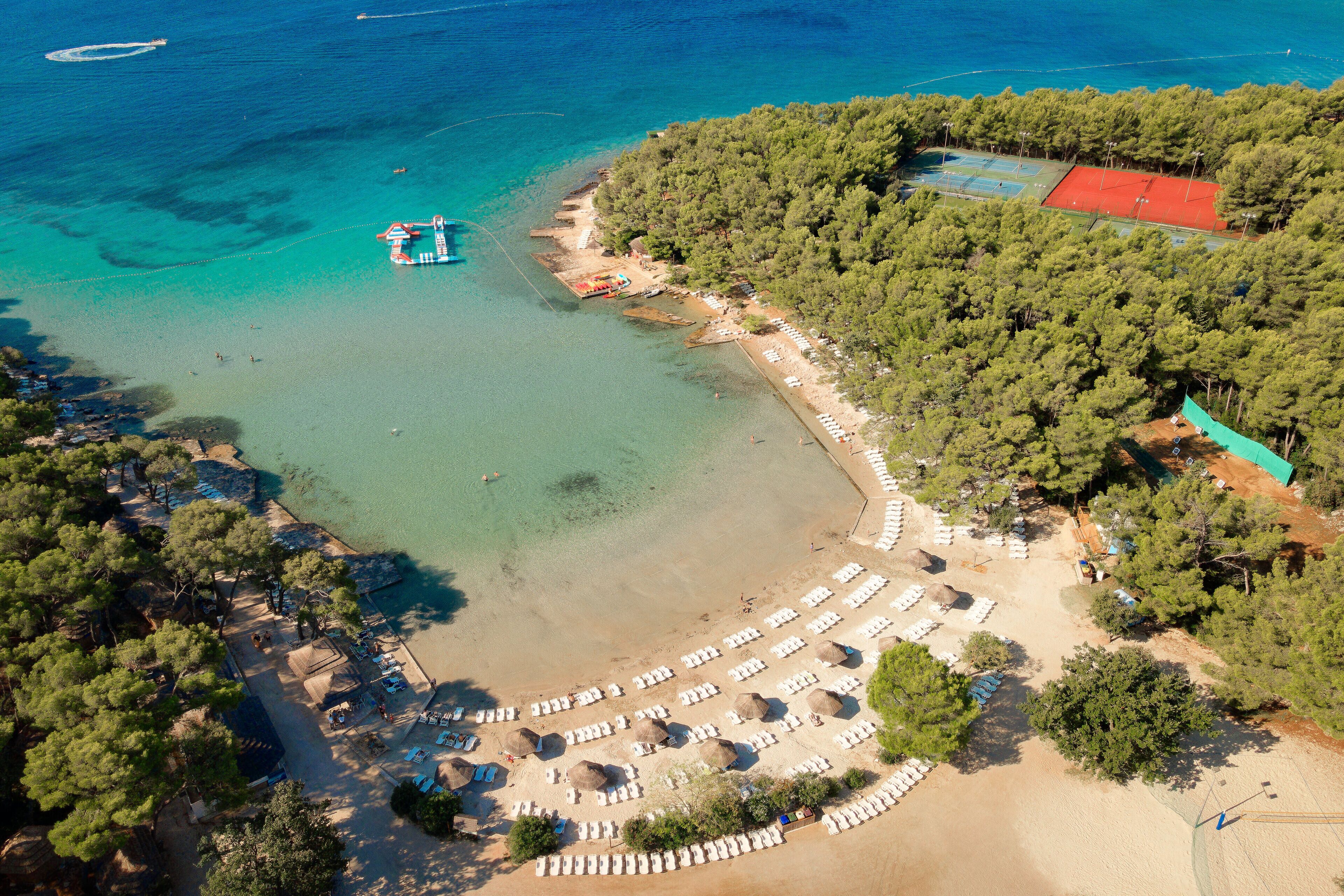An aerial view of a beach with clear turquoise waters, a sandy beach area with rows of sun loungers, and a dense forested area with a tennis court.