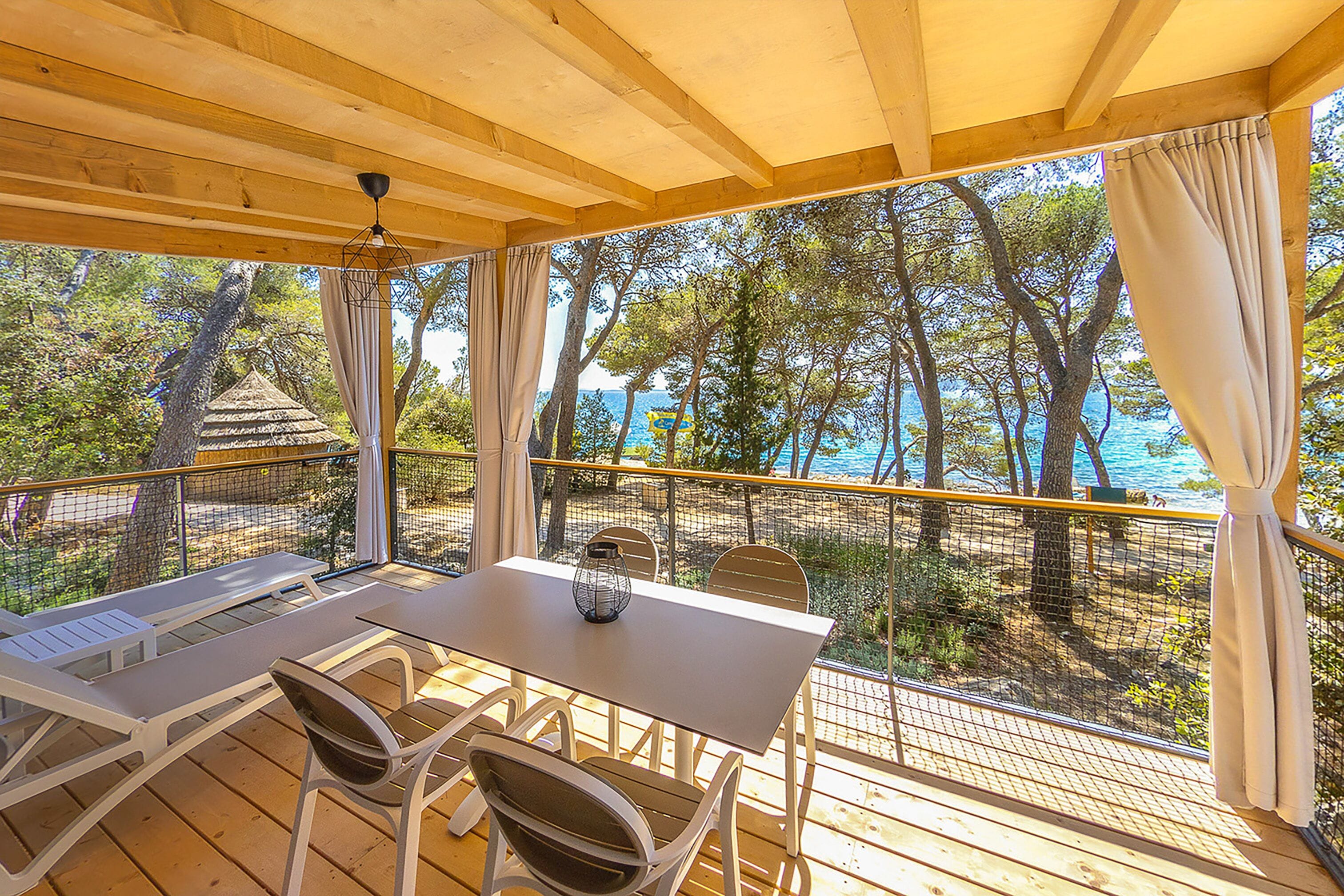 A wooden deck with a table and chairs, overlooking a beach with trees and a clear sky.