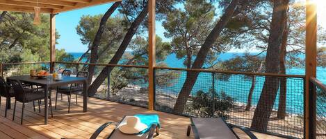 A wooden deck with a table and chairs, overlooking a beach with trees and a clear blue sky.