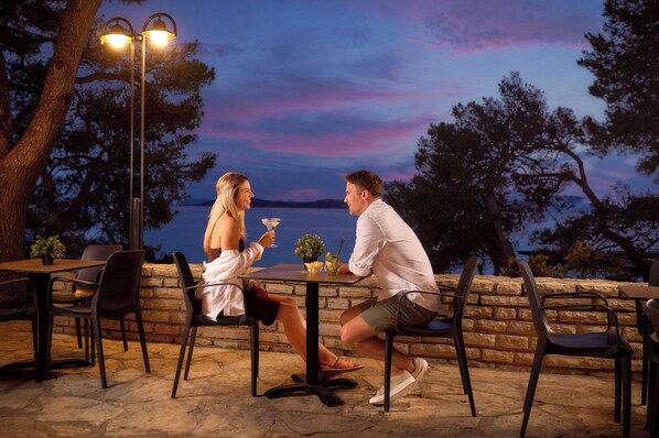 A couple dining at an outdoor table with a view of the water and trees, under a street lamp.
