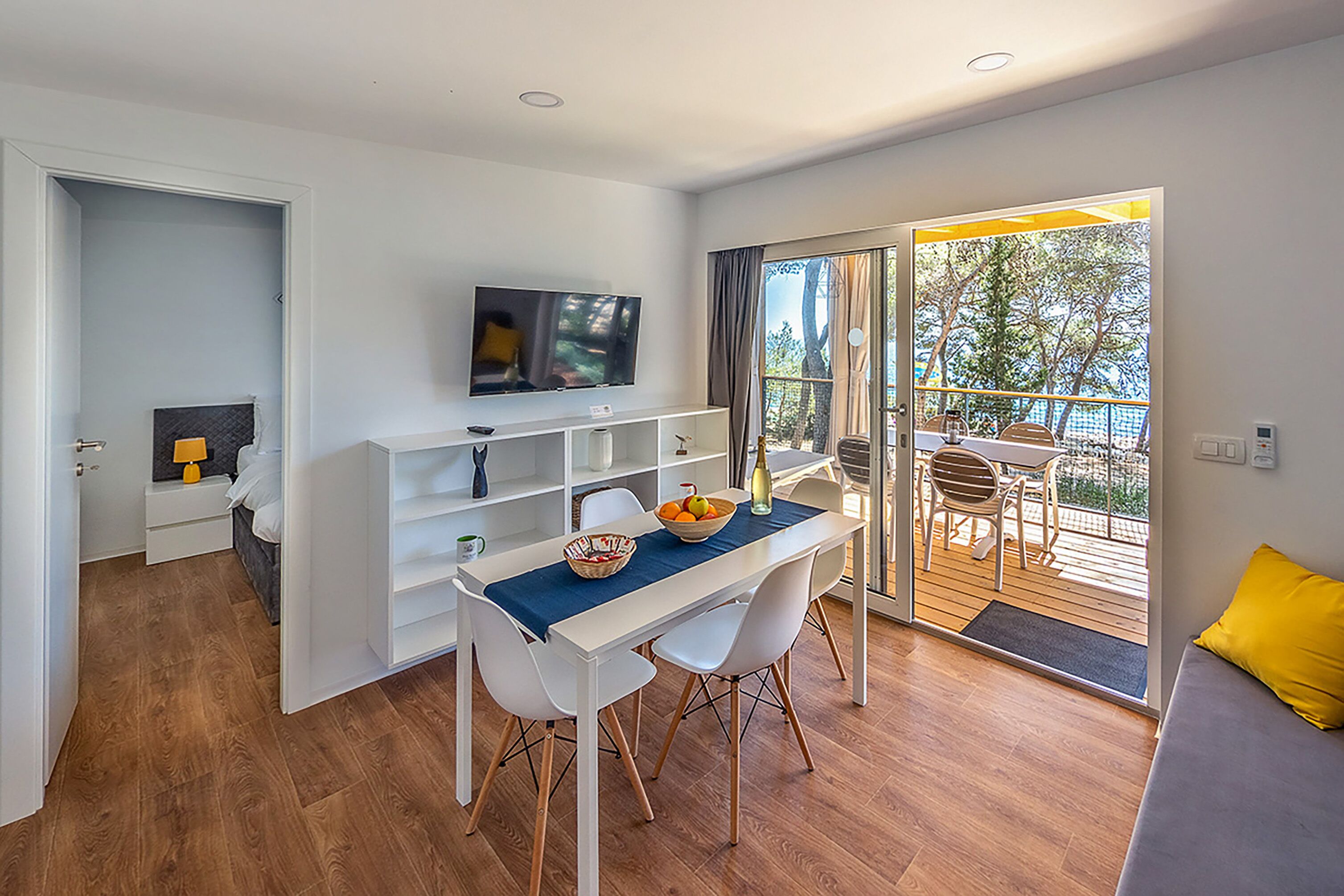 A modern living room with a dining area, a TV mounted on the wall, and a balcony with outdoor furniture.