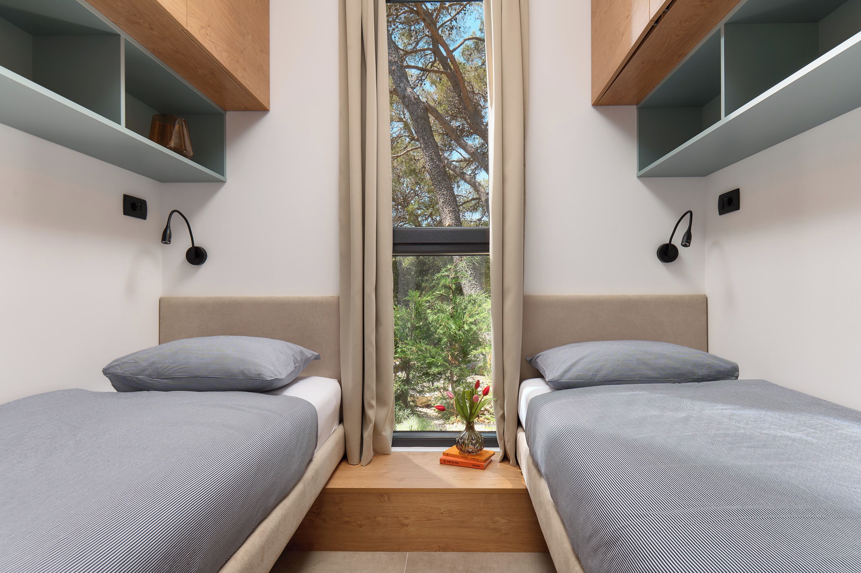 A bedroom with two beds, a window with curtains, a small table with a vase, and built-in shelves.